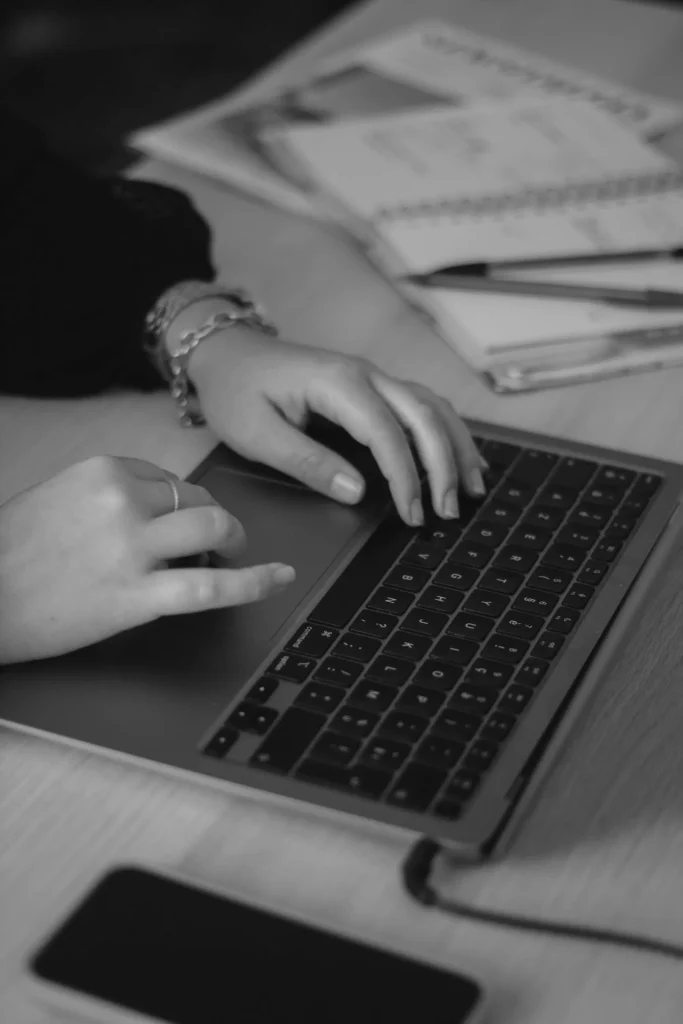 Handen typend op een MacBook-toetsenbord naast een notitieboek en pen tijdens het werken aan een website